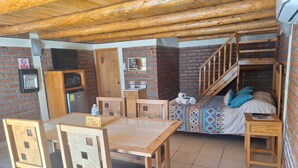 A compact room with a wooden table and chairs, a bed with patterned bedding, a small kitchenette, and a brick wall. - Cabañas Darely (Urique)