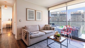A modern living room with a sofa, a coffee table, and a red chair. There are framed documents on the wall and a balcony with a view of buildings. - Apartment - Manquehue - Parque Arauco (santiago)
