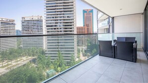 A modern balcony with a glass railing, a wicker sofa, and a view of high-rise buildings and greenery. - Apartment - Manquehue - Parque Arauco (santiago)
