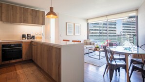 A modern kitchen with wooden cabinets, a built-in oven, and a dining area with a glass table and chairs. - Apartment - Manquehue - Parque Arauco (santiago)