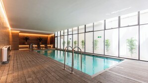 An indoor swimming pool with wooden flooring, metal pool ladders, and large windows offering views of greenery. - Apartment - Manquehue - Parque Arauco (santiago)