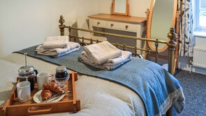 Una cama bien hecha con dos toallas, una bandeja con croissants, café y tazas, un aparador de madera, un espejo y una ventana con cortinas.