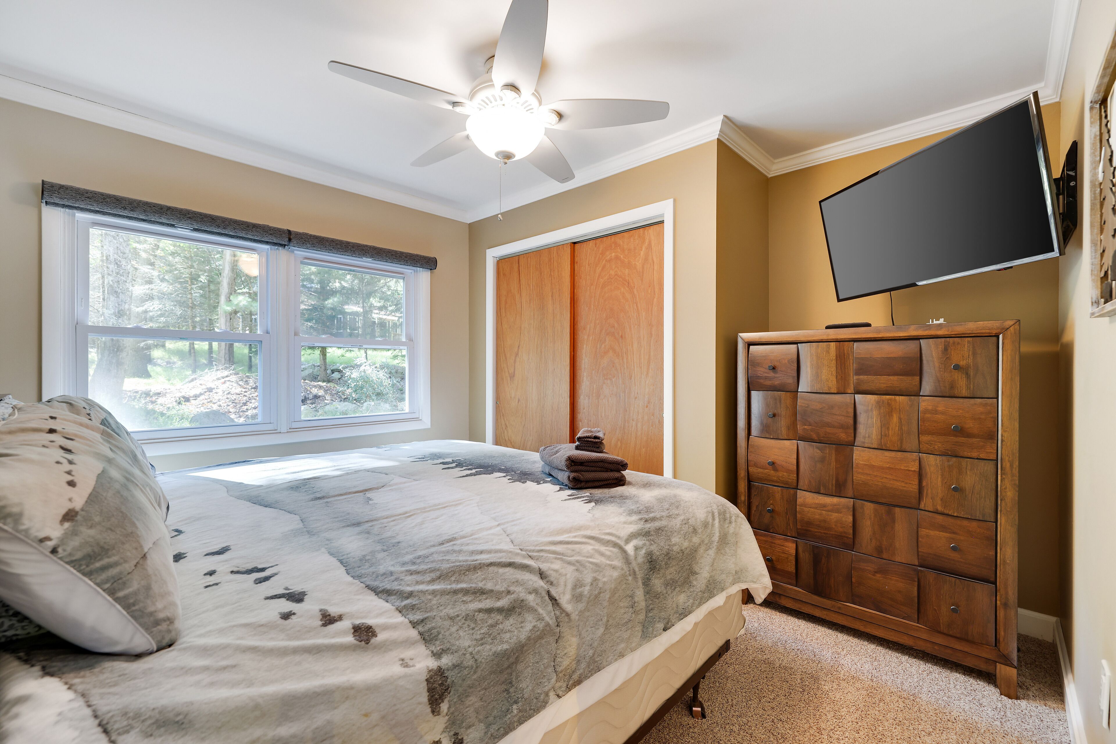 A bedroom with a bed, a wooden dresser, a ceiling fan, a TV, and a window with blinds.