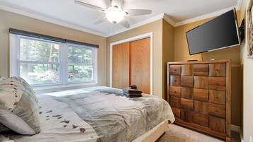 A bedroom with a bed, a wooden dresser, a ceiling fan, a TV, and a window with blinds.