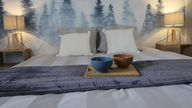 Ein Bett mit grauer Decke, zwei Kissen und ein Holztischchen mit zwei Schüsseln.