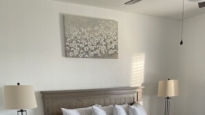 Une chambre à coucher avec un lit en bois, deux tables de chevet, une petite table d’appoint, un ventilateur de plafond et une œuvre d’art accrochée au mur.