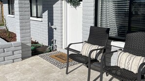 Une terrasse couverte avec deux fauteuils en osier, une porte blanche ornée d’une guirlande et une fenêtre avec des stores.