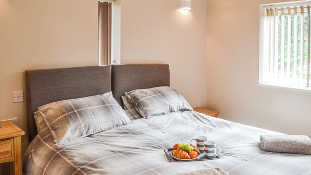Ein ordentlich gemachtes Bett mit einer Karo-Bettdecke, zwei Kissen, einem Tablett mit Croissants und Früchten sowie einem aufgerollten Handtuch.