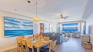 A spacious living area with a dining table set for four, a large window offering a view of the ocean, and a ceiling fan. - UPGRADED-Sands of Kahana #275 OCEANFRONT, 3 Bed Rooms, sleeps 10 - Central AC (Lahaina)