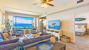 A modern living room with a large window overlooking the ocean, a ceiling fan, and a television. - UPGRADED-Sands of Kahana #275 OCEANFRONT, 3 Bed Rooms, sleeps 10 - Central AC (Lahaina)