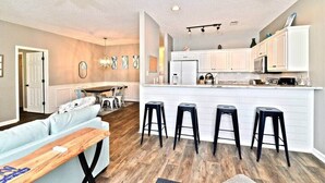 Een moderne keuken met witte kasten, een centraal eiland met krukken, en een zithoek met een lichtblauwe bank en een houten tafel.