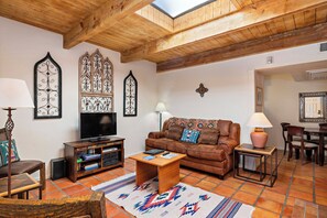 A cozy living room with a wooden ceiling, a brown leather sofa, a TV, and a small table with a lamp. - Fort Marcy 81 | Walk to Plaza | Wood Fireplace | Full Kitchen | Hot Tub | Pool | Single Level (Santa Fe)