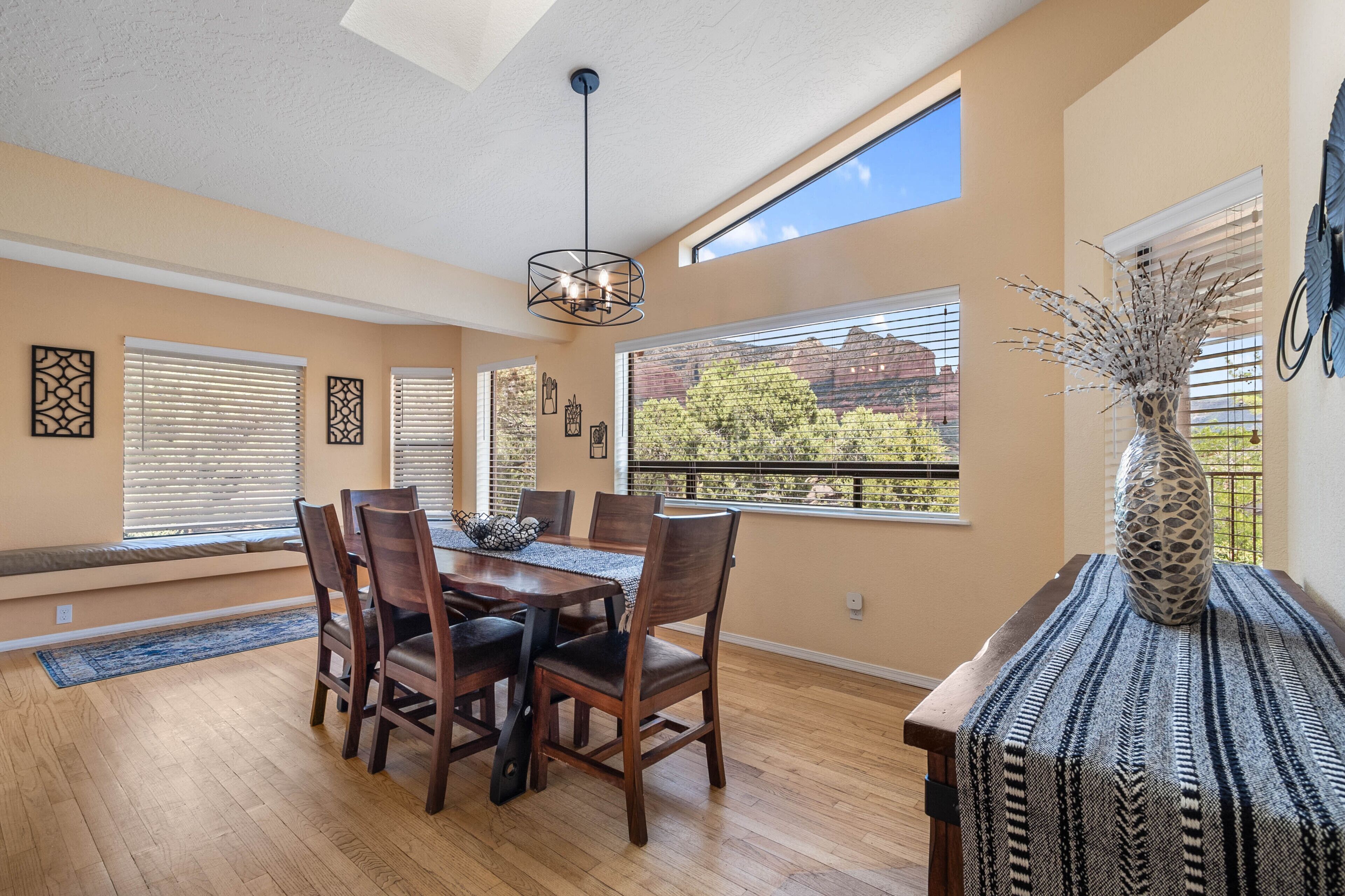 A dining area with a wooden table and chairs, large windows with blinds, and a vase on a side table.