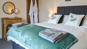 Een net opgemaakt bed met kussens, een nachtkastje met een spiegel, en een deur met daaraan hangende kleding.