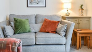 Een woonkamer met een grijze bank, groene en oranje kussens, een lijstwerk, een lamp, een kleine tafel en een kast.
