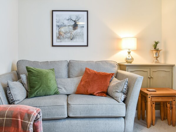 A living room with a grey sofa, green and orange cushions, a framed artwork, a lamp, a small table, and a cabinet.