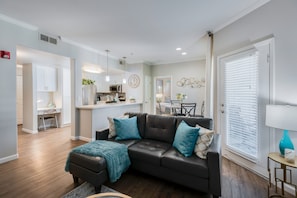 A modern living room with a dark leather sofa, a small coffee table, and a teal throw blanket. There is a kitchen in the background with a microwave and a sink. - King Bed / Prime Location / Private Garage (Roseville)