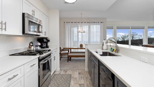 Een moderne keuken met witte kastjes, roestvrijstalen apparatuur, een eethoek met een houten tafel en stoelen, en uitzicht door grote ramen.