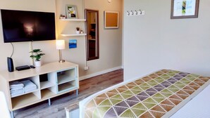 Ein modernes Hotelzimmer mit Flachbildfernseher, Nachttisch mit Lampe, Bett mit Bettdecke mit geometrischem Muster, kleiner Topfpflanze, Regal mit Dekoration und Tür mit Spiegel.