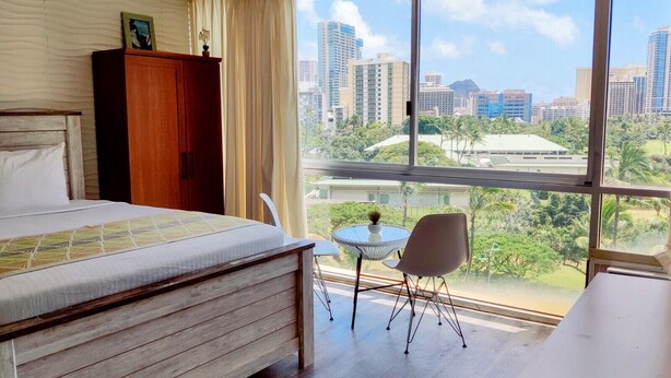Ein Hotelzimmer mit einem großen Bett, einem Holzschrank, einem kleinen runden Tisch mit einem Stuhl und Blick auf eine Stadtlandschaft durch große Fenster.