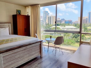 Ein Hotelzimmer mit einem großen Bett, einem Holzschrank, einem kleinen runden Tisch mit einem Stuhl und Blick auf eine Stadtlandschaft durch große Fenster.