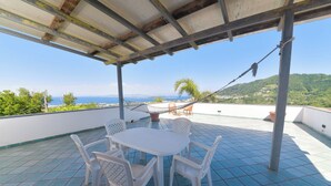 A covered patio with a table and chairs, overlooking a scenic view of the sea and hills. - Villa Stefania - Entire Villa (Barano D'ischia)