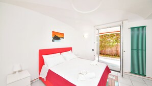 A hotel room with a red bed, white walls, a small bedside table with a lamp, and a view of a garden through an open door. - Villa Stefania - Entire Villa (Barano D'ischia)