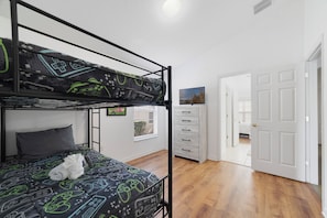 A bunk bed with a gaming-themed bedspread, a dresser, a television, and a door leading to another room. - 4BR Pool Home 15 min to Disney • Heated Pool & Spa • Game Room (Kissimmee)