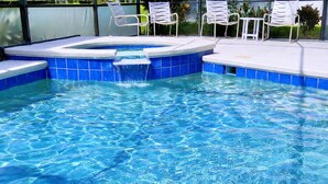 Una piscina con azulejos azules y un jacuzzi. Hay sillas blancas alrededor de la piscina.