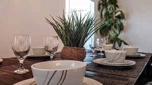 Una mesa de comedor con platos, tazones y copas de vino blancos, una cesta tejida y una planta en maceta.