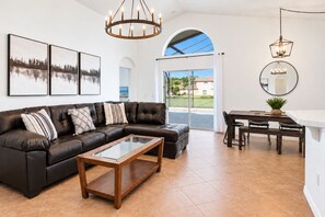 A modern living room with a sectional sofa, a glass coffee table, and a dining area with a table and chairs. - 4BR Pool Home 15 min to Disney • Heated Pool & Spa • Game Room (Kissimmee)
