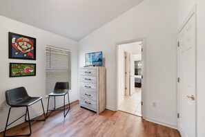 Ein Zimmer mit Holzboden, einem Fernseher auf einem Schrank, einem Sessel, einer Kommode und einer Tür zu einem anderen Raum.