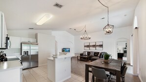 Una cocina moderna con una isla central, un comedor con mesa y sillas de madera, y una sala de estar con sofá y televisor.