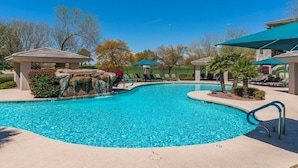 A swimming pool with a curved design, surrounded by lounge chairs and umbrellas, with a stone structure and greenery in the background.