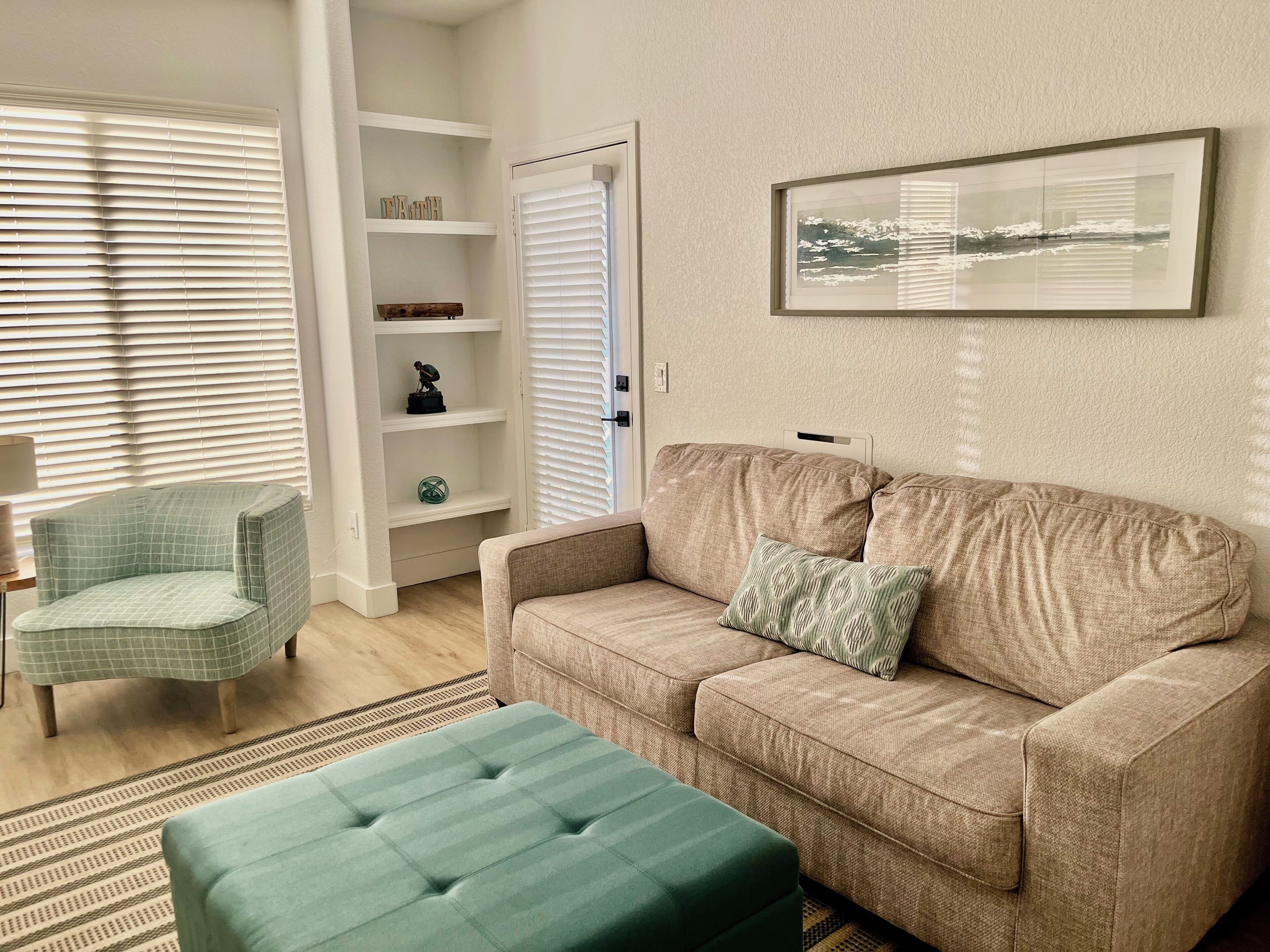 Ett vardagsrum med en beige soffa, en grön pall, en ljusgrön fåtölj och en vit hyllsektion.
