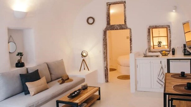 A modern living room with a sofa, coffee table, and a kitchen area with a sink and countertop. - Nykteri Cave | A Terre Cave Villas (Oia)
