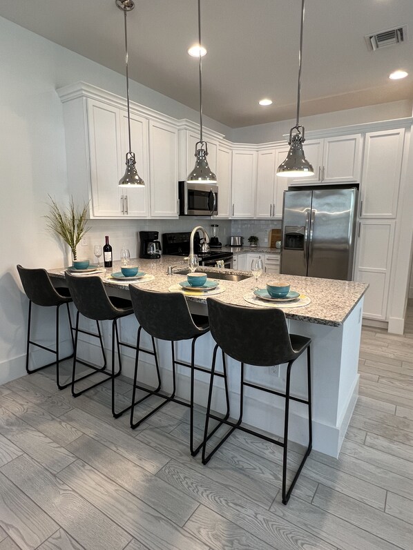 A modern kitchen with white cabinets, a stainless steel refrigerator, a granite countertop, and four bar stools.
