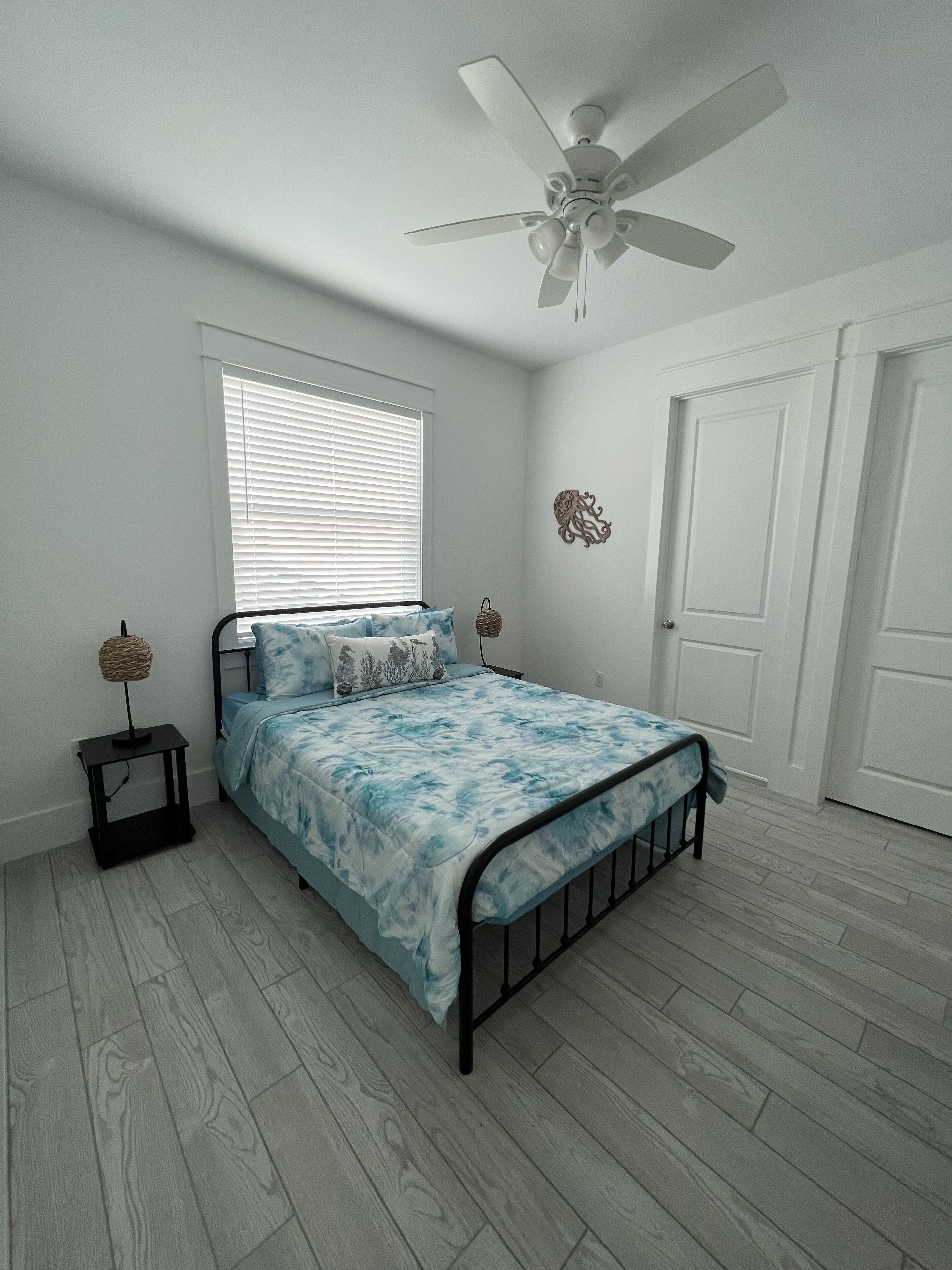 Une chambre à coucher comprenant un lit, un ventilateur de plafond, une fenêtre avec des stores, une porte et une petite table de chevet avec une lampe.