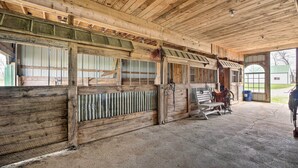 A barn with wooden walls and a concrete floor, featuring stalls with metal gates and a feeding area. - Renovated Bunkhouse on 12-Acre Horse Farm! (Perrysville)