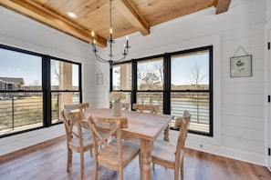 Enjoy meals in this spacious dining area with a large table and six chairs, surrounded by a high ceiling and large windows offering views of the outdoors.