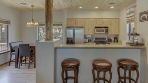 A kitchen with wooden cabinets, a central island with stools, and a dining area with chairs. - Bison Ridge 4 BR, 4BA Cabin with National Forest access and views! (Show Low)