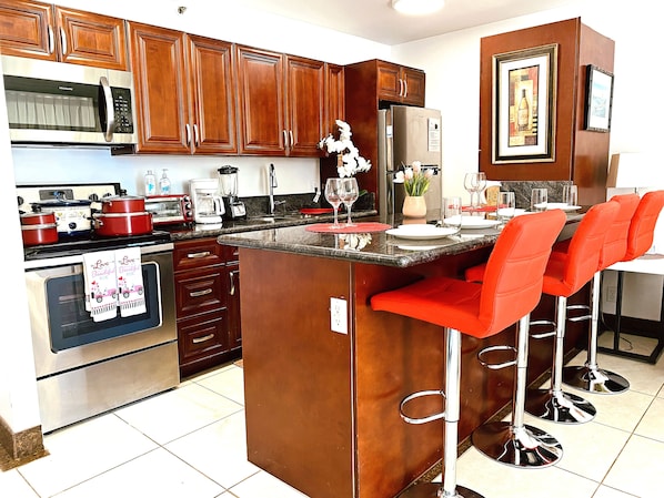 A kitchen with wooden cabinets, stainless steel appliances, a central island with red bar stools, and a dining area with a table set for two. - Waikiki Banyan 1BR - FREE Parking - Walk to Beach & Lanai (Honolulu)