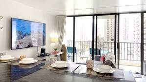 A hotel room with a dining area, a TV, a balcony with a pool view, and a cityscape outside. - Beautiful Waikiki Banyan 1BR/Free PRKG W/ Balcony (Honolulu)