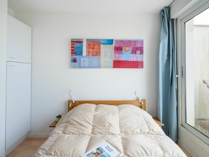 Un dormitorio con cama, una obra de arte en la pared, una ventana con cortinas y una mesita de noche con una planta.