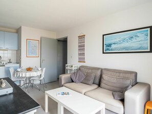 Una sala moderna con un sofá color beige, una mesa de centro blanca y un comedor con una mesa y sillas blancas. Se aprecia una cocina con armarios azul claro y un mapa en la pared.
