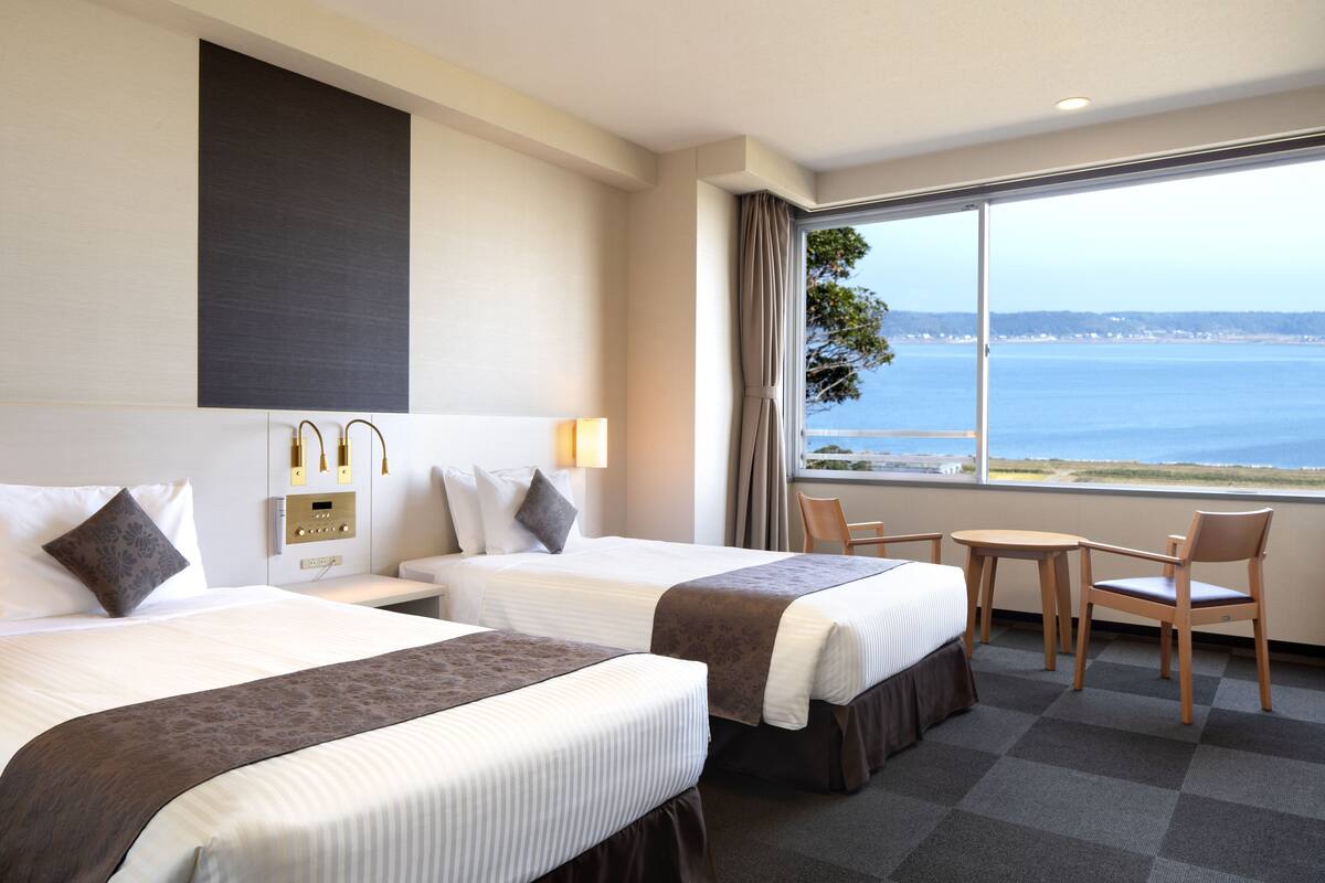 Habitació d'hotel amb dos llits, una taula petita, dues cadires, una finestra amb vista al mar i un panell de control a la paret.