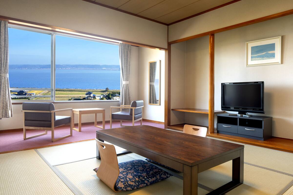 Una habitació amb taula de fusta, dues cadires, televisió i una finestra gran amb vista al mar.