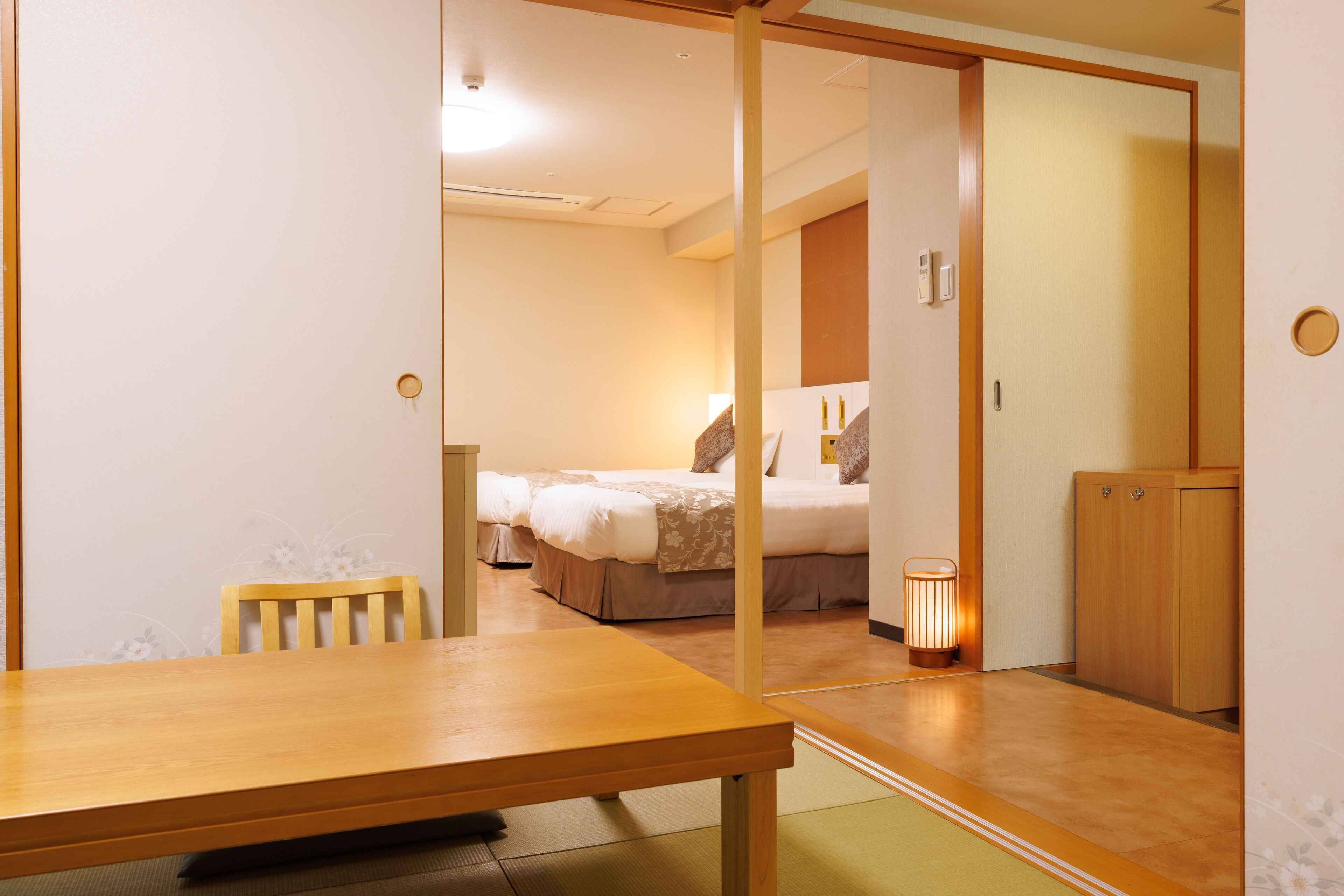 A hotel room with a wooden table, a chair, a bed with pillows, a bedside table with a lamp, and a wardrobe.