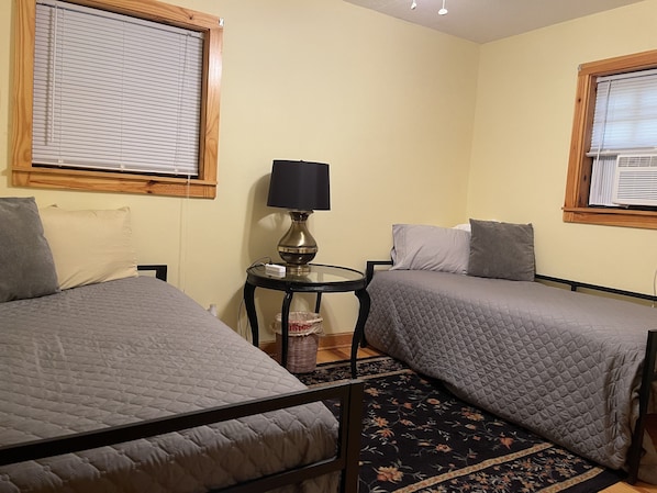 A room with two beds, a small table with a lamp, and a window with blinds.