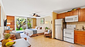 Ein modernes Hotelzimmer mit einem Bett, einer Essecke mit Obstschale, einer Küchenzeile inklusive Kühlschrank und Mikrowelle sowie Gartenblick durch das Fenster.
